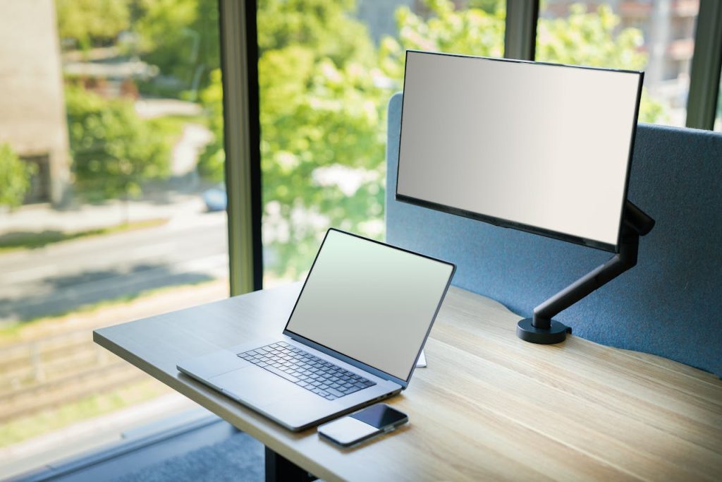 A contemporary office desk setup with a laptop and monitor by a window, featuring bright natural light.